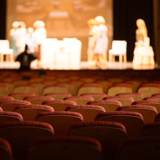Fotografia de uma sala de teatro, da zona do público para o palco, em que vemos atores a encenar uma peça.